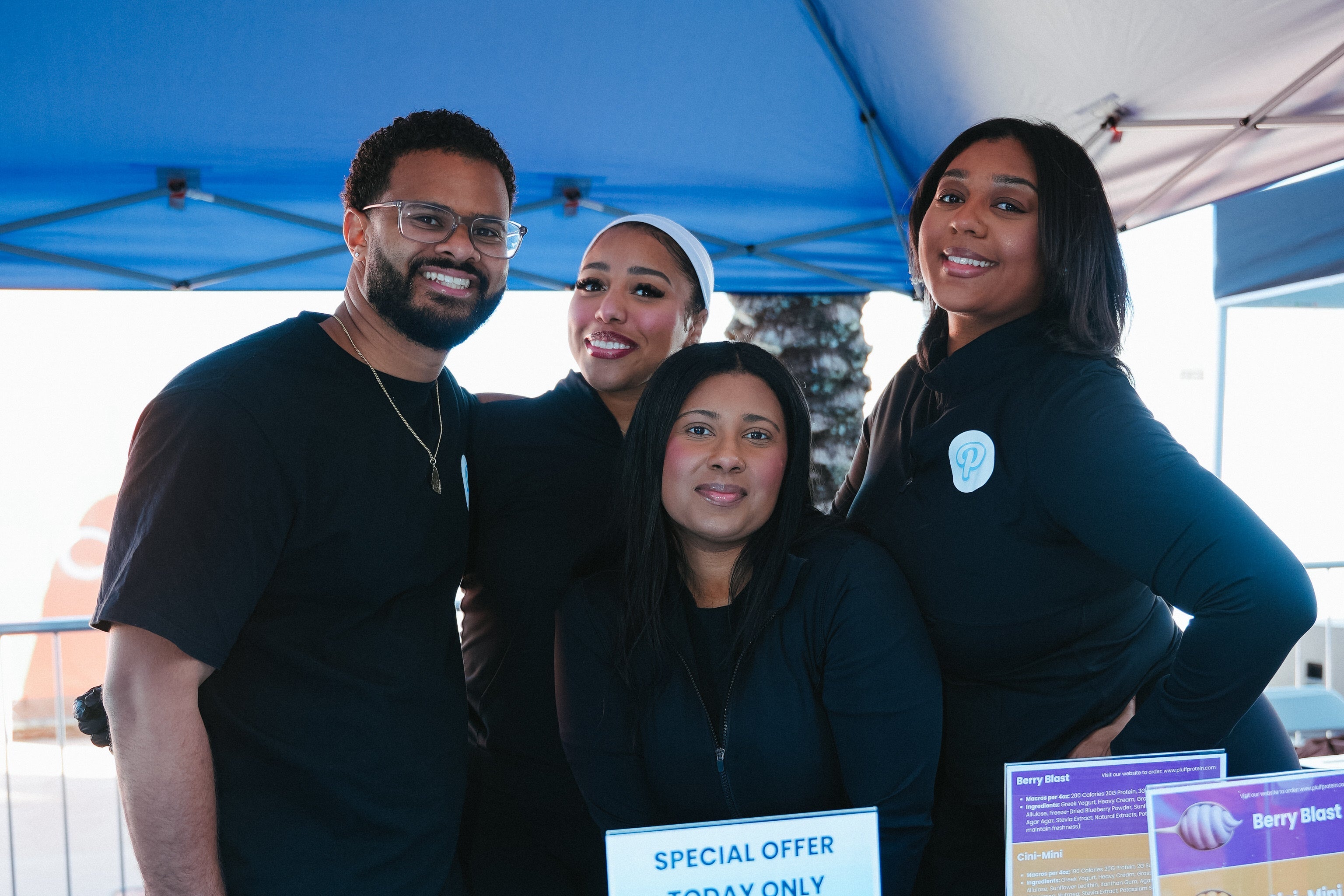 Group of four people standing behind a table with promotional materials under a blue tent.